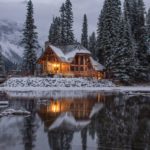 wooden house near pine trees and pond coated with snow during daytime