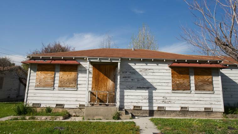 The front part and lawn of an abandoned house with boarded-up windows and door.