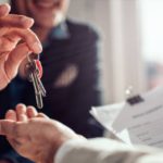 A smiling real estate agent in an office setting handing over house keys to a home buyer holding their lease.