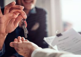 A smiling real estate agent in an office setting handing over house keys to a home buyer holding their lease.