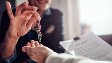 A smiling real estate agent in an office setting handing over house keys to a home buyer holding their lease.