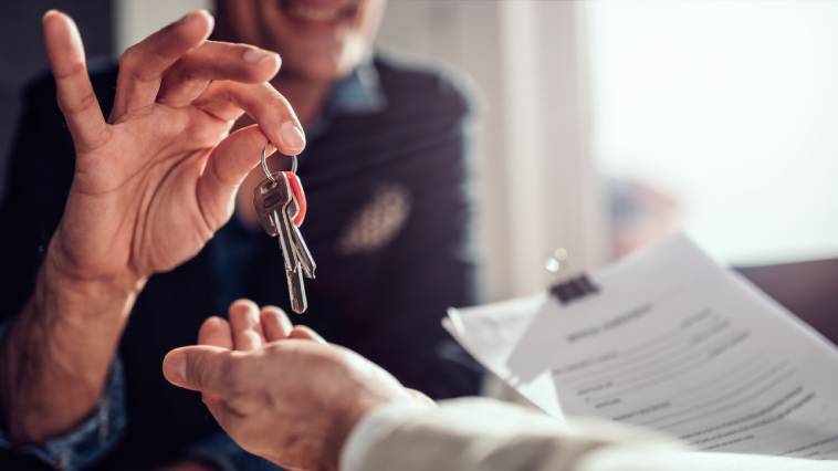 A smiling real estate agent in an office setting handing over house keys to a home buyer holding their lease.