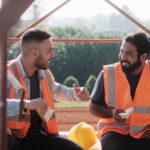 Two happy construction workers wearing orange safety vests talking and laughing while enjoying their lunch break together