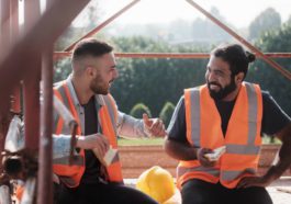 Two happy construction workers wearing orange safety vests talking and laughing while enjoying their lunch break together