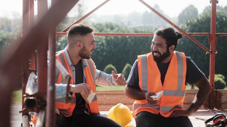 Two happy construction workers wearing orange safety vests talking and laughing while enjoying their lunch break together