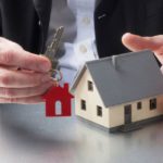 A man in a suit holding the key to a house with a red keychain of a house and a model home in his other hand.