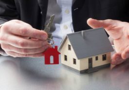 A man in a suit holding the key to a house with a red keychain of a house and a model home in his other hand.