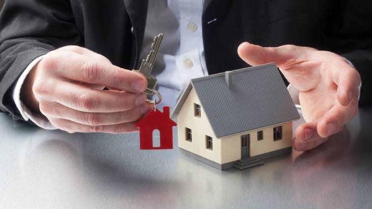 A man in a suit holding the key to a house with a red keychain of a house and a model home in his other hand.